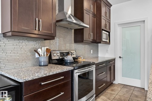 The kitchen features dark wood cabinetry, granite countertops, a tiled backsplash, and stainless steel appliances including a range with an overhead hood fan and a built-in microwave - 26 Edgewater Terr N, St. Albert, AB - Indoor Photo Showing Kitchen With Upgraded Kitchen