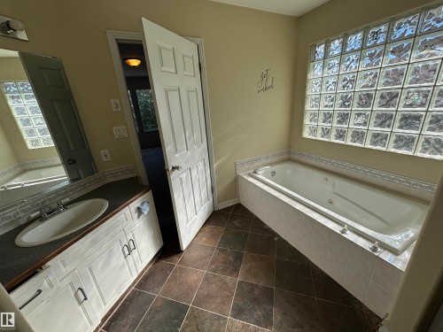 Bathroom featuring a jetted tub, glass block window, dark tile flooring, white vanity with an integrated sink, and an overhead mirror - 3516 49 Avenue, Drayton Valley, AB - Indoor Photo Showing Bathroom