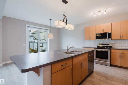 Kitchen featuring a central island with dual sinks, light wood-finish cabinetry, stainless steel appliances, and a subway tile backsplash - 7827 18 Avenue, Edmonton, AB - Indoor Photo Showing Kitchen With Double Sink