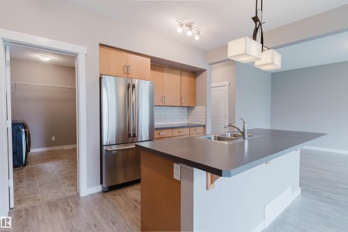 Kitchen featuring wood-finish cabinetry, stainless steel refrigerator, white subway tile backsplash, and dark countertop island with an undermount double-basin sink - 7827 18 Avenue, Edmonton, AB - Indoor Photo Showing Kitchen With Double Sink