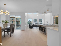 Open-concept living area featuring light wood-finish flooring, a contemporary kitchen island with a waterfall countertop, a dining area with a modern chandelier, and multiple large windows - 