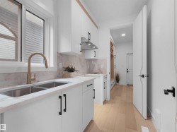 Modern kitchen featuring white cabinetry with matte black hardware, light-toned wood-finish flooring, a double basin stainless steel sink with a brass-finish faucet, white marble-look countertops, and a matching backsplash - 