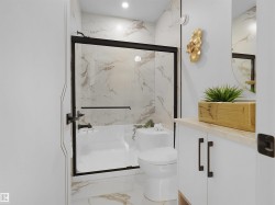 Bathroom featuring a white bathtub with a black-framed glass shower enclosure, a matching toilet, and large format veined tile flooring extending to the shower walls - 