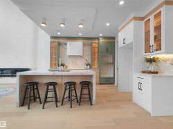 Gourmet kitchen featuring a waterfall-edge island, light wood-finish flooring, custom cabinetry in white and sage tones, integrated range hood, and a stone-patterned backsplash - 