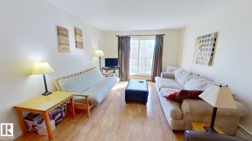 Spacious living area featuring wood-finish flooring, light neutral wall tones, and a large window with vertical blinds and drapery - 401 10326 117 Street, Edmonton, AB - Indoor Photo Showing Living Room