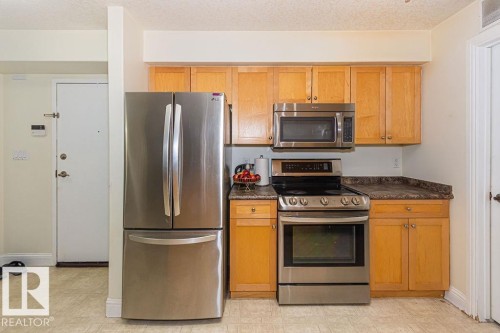 Kitchen featuring light wood cabinetry with brushed nickel hardware, stainless steel appliances including a French door refrigerator and range with overhead microwave, and a dark stone-finish countertop - 214 13005 140 Avenue, Edmonton, AB - Indoor Photo Showing Kitchen