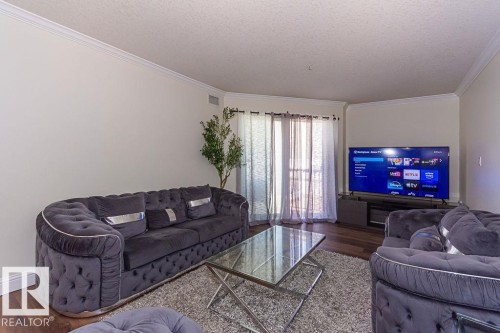 Living area featuring wood-finish flooring, light-toned walls, crown molding, and sliding glass doors with sheer and blackout curtains - 214 13005 140 Avenue, Edmonton, AB - Indoor Photo Showing Living Room