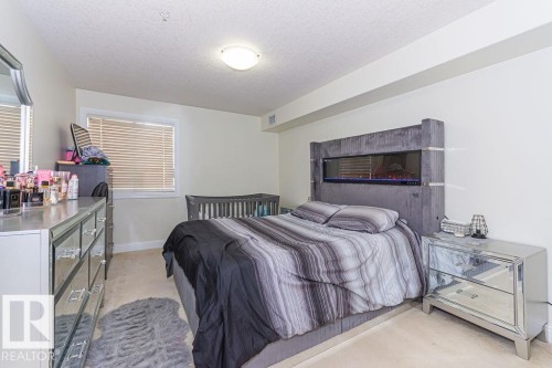 Bright interior space featuring a window with blinds, a flush-mount ceiling light fixture, and a built-in electric fireplace within the headboard - 214 13005 140 Avenue, Edmonton, AB - Indoor Photo Showing Bedroom