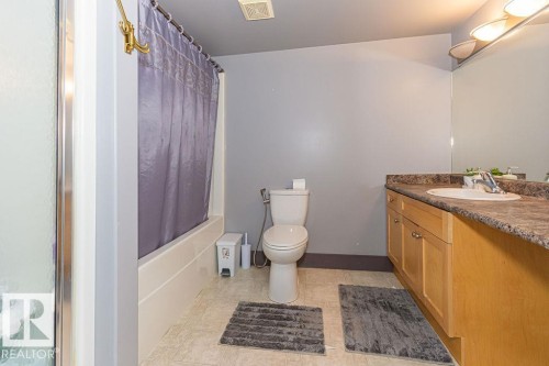 Bathroom featuring a tub/shower combination, light wood-finish vanity with an integrated sink, stone-look countertop, and a wall-mounted mirror - 214 13005 140 Avenue, Edmonton, AB - Indoor Photo Showing Bathroom