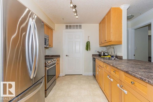 Galley kitchen featuring stainless steel appliances, light wood-finish cabinetry, and speckled countertops - 214 13005 140 Avenue, Edmonton, AB - Indoor Photo Showing Kitchen With Double Sink