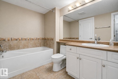 Bathroom featuring a white vanity with built-in sink, a large wall-mounted mirror, and a full bathtub with a tiled surround - 204 278 Suder Greens Drive, Edmonton, AB - Indoor Photo Showing Bathroom