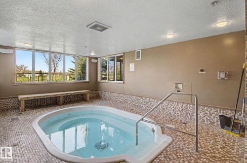 Indoor hot tub featuring a stainless steel handrail, mosaic tile surround, and large windows - 204 278 Suder Greens Drive, Edmonton, AB - Indoor Photo Showing Other Room