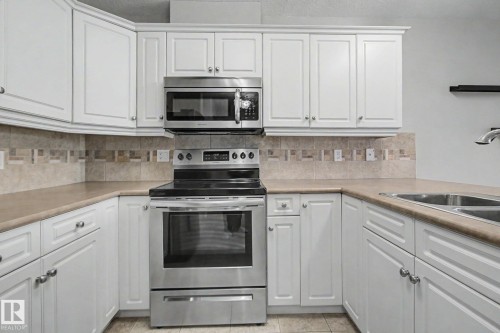 Bright kitchen featuring white cabinetry, stainless steel appliances, a tiled backsplash, and double basin sink - 204 278 Suder Greens Drive, Edmonton, AB - Indoor Photo Showing Kitchen