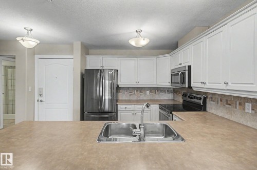Kitchen featuring a dual basin stainless steel sink, expansive countertop, white cabinetry with crown molding, stainless steel appliances, and a tiled backsplash - 204 278 Suder Greens Drive, Edmonton, AB - Indoor Photo Showing Kitchen With Double Sink