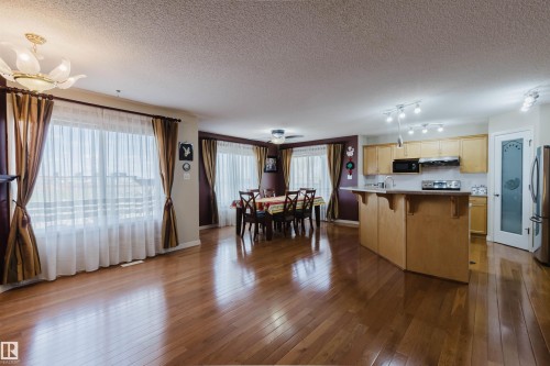 Open concept living space featuring wood-finish flooring, a kitchen with light wood cabinetry and a central island, a dedicated dining area, and multiple large windows with sheer and opaque drapery - 1764 Melrose Crescent, Edmonton, AB 