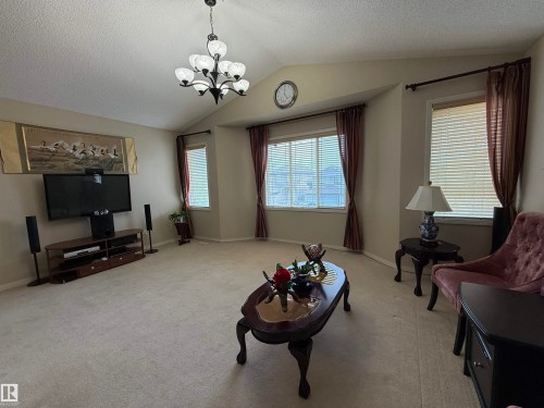 Spacious living area featuring a vaulted ceiling with a central chandelier, multiple windows with blinds and drapes, and neutral wall tones - 1764 Melrose Crescent, Edmonton, AB 