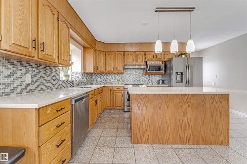 Kitchen featuring wood-finish cabinetry, stainless steel appliances, a mosaic tile backsplash, light-toned countertops, and an island with pendant lighting - 1529 49A Street Nw, Edmonton, AB - Indoor Photo Showing Kitchen With Upgraded Kitchen