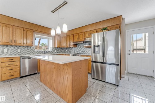 Kitchen featuring a central island with a white countertop, wood-finish cabinetry, stainless steel appliances, patterned tile backsplash, and light-toned tile flooring - 1529 49A Street Nw, Edmonton, AB - Indoor Photo Showing Kitchen With Upgraded Kitchen