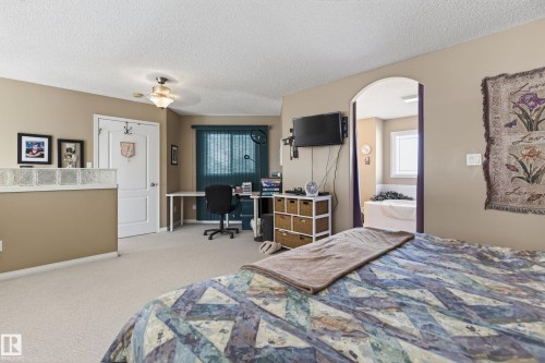 Carpeted room featuring an arched doorway leading to a space with a built-in tub, a wall-mounted light fixture, and a window with blinds - 21 Pipestone Drive, Devon, AB - Indoor Photo Showing Bedroom