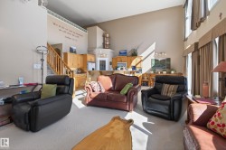 Open-concept living area with vaulted ceilings, large windows with vertical blinds, light-toned carpeting, and a built-in kitchen pass-through with wood cabinetry - 