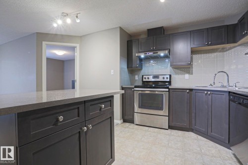 Modern kitchen featuring dark cabinetry with silver hardware, light-toned countertops, and a stainless steel range with overhead ventilation - 201 1520 Hammond Gate Gate, Edmonton, AB 