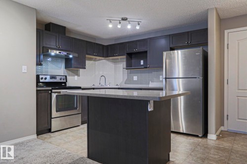 Contemporary kitchen featuring dark cabinetry, stainless steel appliances, a light-toned backsplash, a central island with a solid surface countertop, and track lighting - 201 1520 Hammond Gate Gate, Edmonton, AB 