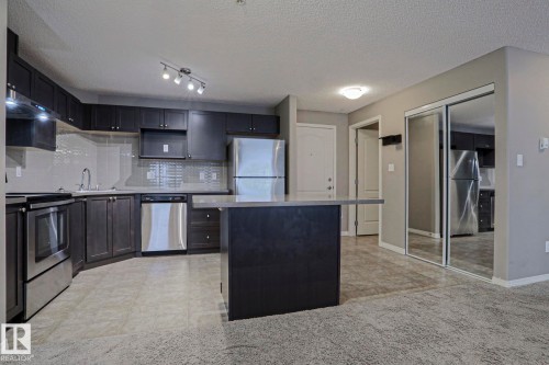 Kitchen featuring dark wood-finish cabinetry, stainless steel appliances, a central island with a solid surface countertop, and a tiled backsplash - 201 1520 Hammond Gate Gate, Edmonton, AB 