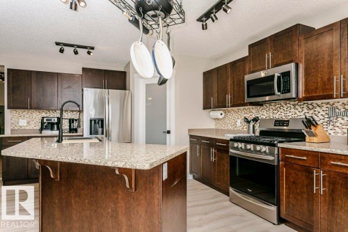 Kitchen featuring a central island with a sink, dark wood-finish cabinetry, stainless steel appliances, speckled countertops, and a mosaic tile backsplash - 42 Santana Crescent, Fort Saskatchewan, AB - Indoor Photo Showing Kitchen With Stainless Steel Kitchen With Upgraded Kitchen