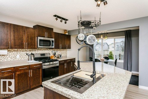Kitchen featuring an expansive island with an undermount sink, granite-style countertops, wood-finish cabinetry, stainless steel appliances, and a mosaic tile backsplash - 42 Santana Crescent, Fort Saskatchewan, AB - Indoor Photo Showing Kitchen With Stainless Steel Kitchen With Double Sink With Upgraded Kitchen