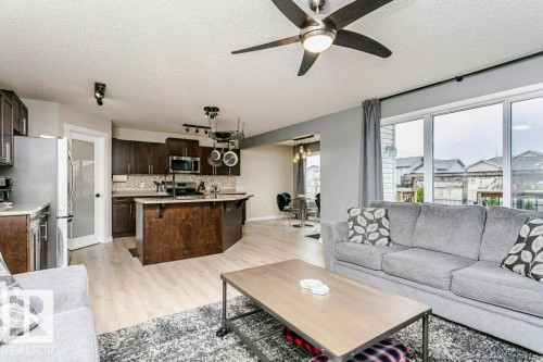 Open-concept living space with light wood-finish flooring, a kitchen featuring dark wood cabinetry and stainless steel appliances, a kitchen island with seating, a ceiling fan, and large sliding glass doors providing access to the exterior - 42 Santana Crescent, Fort Saskatchewan, AB - Indoor Photo Showing Living Room