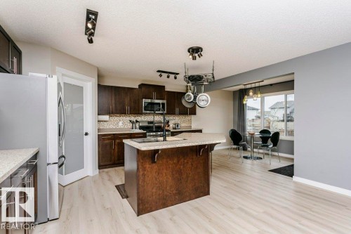 Open-concept kitchen featuring dark wood cabinetry, light granite-finish countertops, a central island with integrated sink, stainless steel appliances, and wood-finish flooring - 42 Santana Crescent, Fort Saskatchewan, AB - Indoor Photo Showing Kitchen