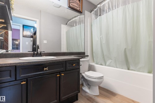Bathroom featuring a dark wood-finish vanity with a single sink and matte black faucet, a full-width mirror, a white bathtub with shower, and wood-finish flooring - 73 3040 Spence Wynd, Edmonton, AB - Indoor Photo Showing Bathroom