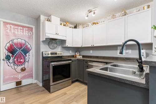Kitchen featuring two-tone cabinetry with white upper cabinets and gray lower cabinets, stainless steel appliances, a double basin sink with a matte black faucet, and wood-finish flooring - 73 3040 Spence Wynd, Edmonton, AB - Indoor Photo Showing Kitchen With Double Sink