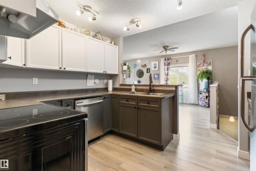 Kitchen featuring an electric range, stainless steel dishwasher, dual-tone cabinetry, dark countertops, and wood-finish flooring - 73 3040 Spence Wynd, Edmonton, AB - Indoor Photo Showing Kitchen With Stainless Steel Kitchen With Double Sink
