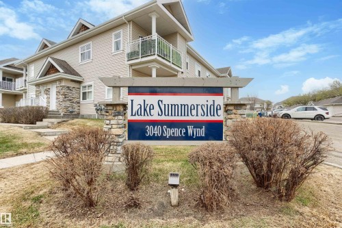 Stone-clad entry sign featuring "Lake Summerside 3040 Spence Wynd" lettering, with a multi-story building exhibiting light-toned siding, white-railed balconies, and stone accents - 73 3040 Spence Wynd, Edmonton, AB - Outdoor