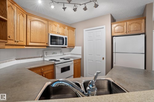 Kitchen featuring wood cabinetry, laminate countertops, a double basin stainless steel sink with a chrome faucet, white tile backsplash, and white appliances including a refrigerator, range, and microwave - 104 9760 174 Street, Edmonton, AB - Indoor Photo Showing Kitchen With Double Sink