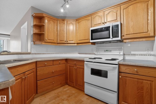 Corner kitchen featuring wood-finish cabinetry, solid surface countertops, a white tile backsplash with decorative trim, and wood-finish flooring - 104 9760 174 Street, Edmonton, AB - Indoor Photo Showing Kitchen