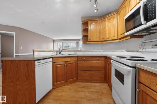 Kitchen featuring wood cabinetry, light-toned countertops, a built-in dishwasher, white appliances, and wood-finish flooring - 104 9760 174 Street, Edmonton, AB - Indoor Photo Showing Kitchen