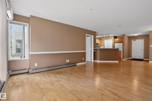 Open-concept living area featuring light wood-finish flooring, warm-toned walls with chair rail molding, and a window with a split-system air conditioner - 104 9760 174 Street, Edmonton, AB - Indoor Photo Showing Other Room