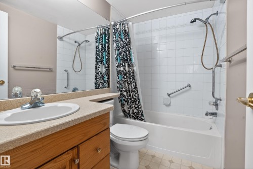 Full bathroom featuring a white bathtub with a tiled surround, a vanity with a light-toned countertop and integrated sink, a large wall-mounted mirror, and wood-finish cabinetry - 104 9760 174 Street, Edmonton, AB - Indoor Photo Showing Bathroom