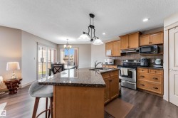 The kitchen features wood cabinetry, stainless steel appliances, a kitchen island with a granite countertop, and dark flooring - 