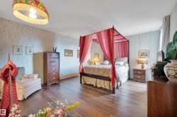 Spacious bedroom featuring wood-finish flooring, patterned wallpaper, a decorative ceiling light fixture, and window with drapes - 