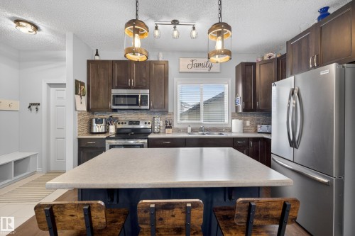 Kitchen featuring a central island with countertop seating, dark wood cabinetry, stainless steel appliances, mosaic tile backsplash, and overhead pendant lighting - 7074 Cardinal Way, Edmonton, AB - Indoor Photo Showing Kitchen With Double Sink