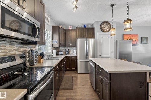 Kitchen featuring wood-finish flooring, stainless steel appliances, dark wood cabinetry, a mosaic tile backsplash, and a central island with a light-toned countertop - 7074 Cardinal Way, Edmonton, AB - Indoor Photo Showing Kitchen With Double Sink