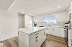 Modern kitchen featuring a central island with white countertop, wood-finish flooring, white cabinetry with bronze hardware, and recessed lighting - 