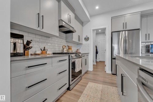 Modern kitchen featuring light gray cabinetry with black bar pulls, subway tile backsplash, stainless steel appliances, light-toned countertops, and wood-finish flooring - 17319 2 St Nw, Edmonton, AB - Indoor Photo Showing Kitchen With Stainless Steel Kitchen With Upgraded Kitchen