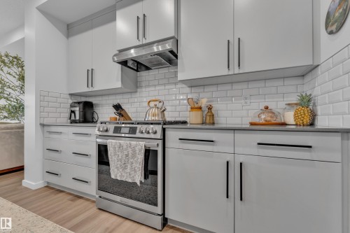 Modern kitchen featuring light gray cabinetry with black bar pulls, a stainless steel range and hood, white subway tile backsplash, dark countertops, and wood-finish flooring - 17319 2 St Nw, Edmonton, AB - Indoor Photo Showing Kitchen With Upgraded Kitchen