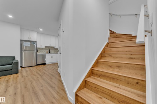 Wood-finish staircase with a white handrail, white walls, recessed lighting, and wood-finish flooring extending into the kitchen featuring white cabinetry, stainless steel appliances, and a tiled backsplash - 17319 2 St Nw, Edmonton, AB - Indoor