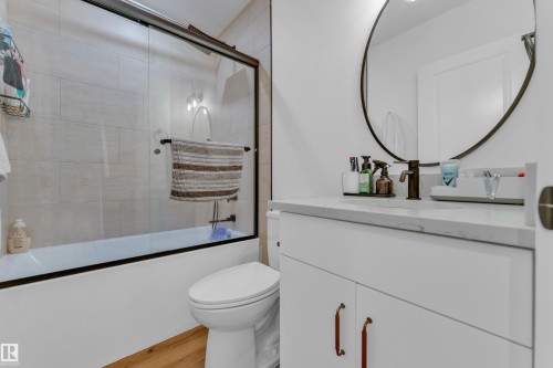 Bathroom featuring a white vanity with a stone-finish countertop, a circular mirror, and a tub with a sliding glass door - 17319 2 St Nw, Edmonton, AB - Indoor Photo Showing Bathroom