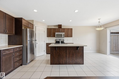 Functional kitchen featuring a central island with a sink, stainless steel appliances, dark wood-finish cabinetry, light countertops, and white square tile flooring - 2170 52A Street Sw, Edmonton, AB - Indoor Photo Showing Kitchen With Stainless Steel Kitchen